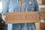 A person holding a paper sign that says “volunteers needed”.