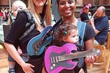 White woman and black woman carrying babies and foam guitars. Other people in the background