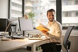 A man seated in front of a naturally lit office desk, smiling.