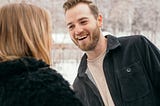 The image shows a woman from behind talking to a man who is facing her. We can see that he is making eye contact with her and looks like he may be chuckling at something she said.