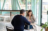 A man and a woman sit at a table and look together at the screen of a laptop.