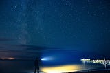 A person with a flashlight looking out over a dark ocean and the starry sky