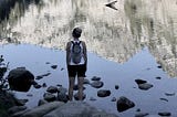 A girl standing in front of an alpine lake, her back to the camera, looking into the distance probably pondering who she is and the meaning of life