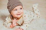 A smiling baby lying on a soft white fur rug. The infant is wearing a brown and white striped beanie hat and a light beige onesie with small brown and orange patterns. The baby is looking up at the camera with a happy expression and has one hand raised as if waving. The setting appears to be a home with wooden flooring visible in the background.