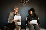 Two women sitting on chairs looking at papers. The left one is older, wearing a business suit, and looks more professional, but she’s still listening attentively to the younger one