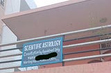 An image shows a hand-lettered sign with a blue background and white text mounted on a railing of a building. The building has a light brown exterior and a pink overhang. The sign displays the following text: “SCIENTIFIC ASTROLOGY”, “Consultation by Appointment Only”, “Mobile” (with a blacked-out section), and “Working Hours: 11.30 AM to 1.30 PM”. In the background, parts of other buildings and some greenery are visible.