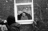 A monochrome picture. An elderly woman with glasses looks out of the window of a stone house. Next to the window hangs a plant pot in the shape of a cornucopia. Vines grow out of it. A group of people dressed like youngsters is passing by the window, their faces can’t be seen.
