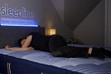 A person is lying on a bed. He appears to be doing a trial of a mattress or brand of bed since the brand is shown on a lighted sign above the bed.
