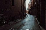 A curving pathway slick with rain, surrounding by imposing buildings. It’s daylight, but all is dark and in shadow, except for a bright sky in the background. In the foreground is the word stop written in large white letters on the narrow road. The word is upside down.