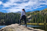 Me standing on a rock overlooking a lake with alpine trees and mountain tops in the distance