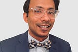 Headshot of Ather, who is Asian, smiling, wearing a navy suit and checkered bowtie. He is wearing thick clear-framed glasses.