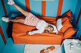 Girl awkwardly lying on orange bed with her feet on the blue and orange striped wall in a tiny room
