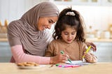 A woman wearing a Hijab helping a schoolchild (possibly with autism or Asperger’s) with her work.