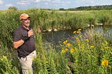 a man with a microphone stands at the edge of a pond surrounded by tall flowers and grasses