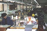 Two rows of industrial sewing machines extending backwards in a factory in China. The first two machines are being operated by a man and a woman who are smiling as they work.