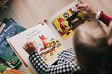 A child and an adult are reading a colorful children’s book together. The page shows an illustration of a red fox wearing trousers, looking for his socks in a room with scattered items. The text on the page reads, “Poor old Fox has lost his socks.”