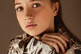 Young teenage girl with brown hair and light eyes stares at the Photographer Head shot. Girl is not smelling, and wears an artistic brown and white jacket.