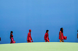 Models wearing red outfits walking across a green hill in a line with a blue backdrop.