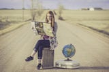 A young woman traveler sitting on a wood crate next to a globe on top of a suitcase. She’s in the middle of the road and holding a map.