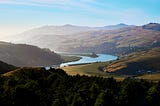 Long distance vista of golden mountain ridges below blue sky, towering over a blue winding sinuous river, through a distant valley with a small village, surrounded by forest at dusk.