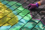 Close up image of a hand tracing a chalk outline around a colourful pattern on a brick pavement.