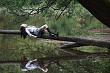 woman, with an open book covering her face, lying on tree trunk that leans out over a reflective lake