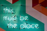 Neon sign on a colorblock wall that reads “this must be the place”