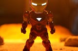 A superhero toy in a “superhero pose” — feet apart, arms ready for action — with bright eyes in front of a dim yellowish light.