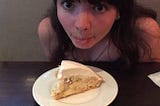 Anna leans head over banana cream pie on plate, sticking tongue out and making wide googly eyes at it.