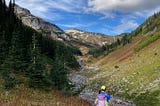 A hiker faces towards the head of a beautful valley with fir trees and ground foliage turning fall colors, blue sky with a few whispy clouds