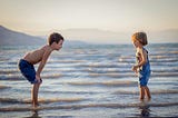 Two boys stare at each other on a beach, their feet in the water.
