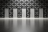 Black and white photo of a wall with decorative wallpaper. On the wall are five white doors. In the foreground of the photo are bare wood floors.