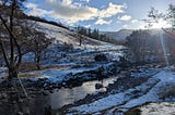 A shallow stream flows through a snowy hillside landscape with bare trees and distant hills under a blue sky.