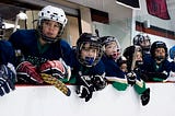 Hockey players lean against the boards to watch a game in progress. They are all wearing the same blue, white and green jerseys.