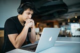 Image of a young man wearing black wireless headphones and is deeply focused on his macbook that is in front of him.