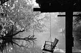 A porch swing hangs from the roof of a ramshackle dock on a river. In the distance are trees whose branches extend out over the river. The air appears foggy or misty. The photo is in black and white.