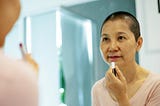 A middle aged woman with a shaved head applying lipstick in the mirror