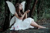 A beautiful dark-haired woman dressed in a white fairy costume sits in the forest holding colorful butterflies
