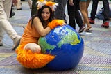 A woman in an orange-eared and cuffed Firefox cosplay hugging an inflatable globe, smiling as she sits on a patterned floor.