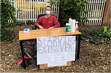 A young man sitting at a desk with a sign that reads, “Starving Extrovert.”