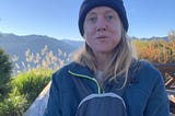 A picture of a blonder woman eating a snack, dressed warmly on a mountain in the sun