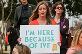 Picture of a woman looking at the camera holding a sign that reads “I’M HERE BECAUSE OF IVF”