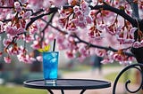 Image shows an overhanging branch with beautiful budding blossoms, and the top of a bistro table with bright blue cocktail in a glass on it.