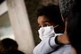 Small child with dark skin and curly black hair wears a face mask while being carried by an adult. The child looks over the adult’s shoulder, clinging to the adult, eyes wide.