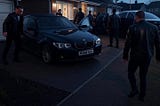 A squad of men in black leather jackets swarm a quiet suburban driveway in the pre-dawn light, moments before pounding on John Morton’s door.