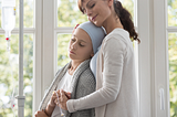 Mother comforting her daughter who is hooked up to an iv and has a head covering.