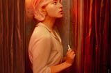 A digital painting of Eddie, a young blonde wearing a shirt and trousers, with an anxious expression. She’s holding a gold key as she stands in a small booth lit by a red bulb.