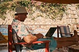 A man wearing an Hawaiian shirt sitting poolside with a compter on his lap and a drink by his side.