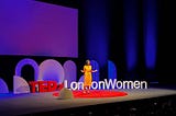 Anneka on stage wearing yellow dress, with arms opened out. Standing on big red circular carpet on a black stage. Behind are the letters ‘TEDxLondonWomen’ against purple-blue stage lights.