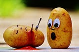 A potato with a drawn on face has been killed by a tiny knife and fork. It is bleeding out ketchup. Another potato looks on, shocked.